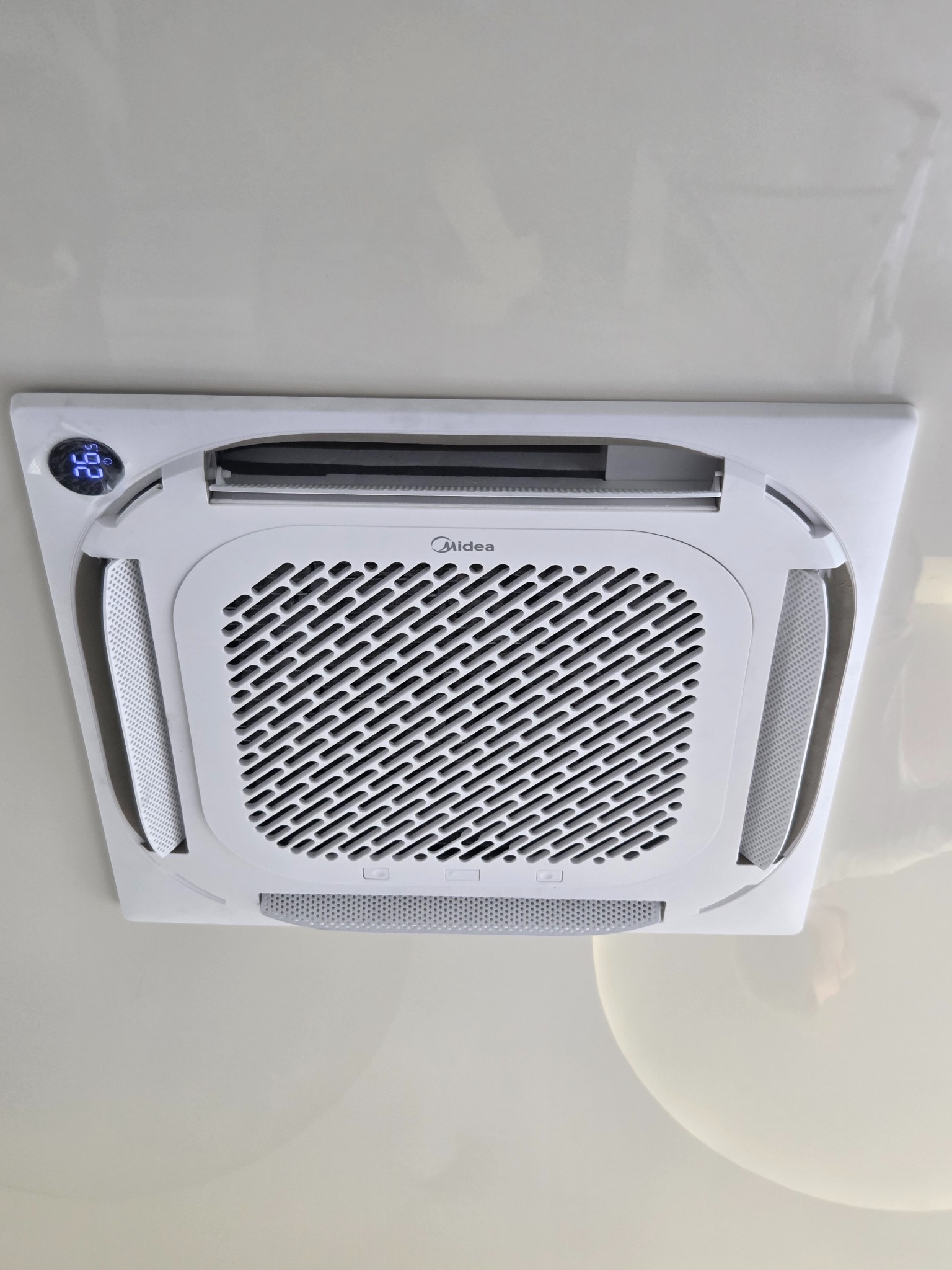 Biała, sufitowa klimatyzacja Midea z wyświetlaczem temperatury 26.5°C, zamontowana na jasnym suficie. Widok od dołu.