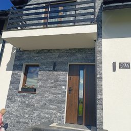 FIRMA REMONTOWO-WYKOŃCZENIOWA ROMAN ZAJĄC - Elewacja domu z szarej cegły, balkon z czarną balustradą, brązowe drzwi wejściowe z przeszkleniem, numer domu 596, małe okno z kwiatami. Błękitne niebo w tle.