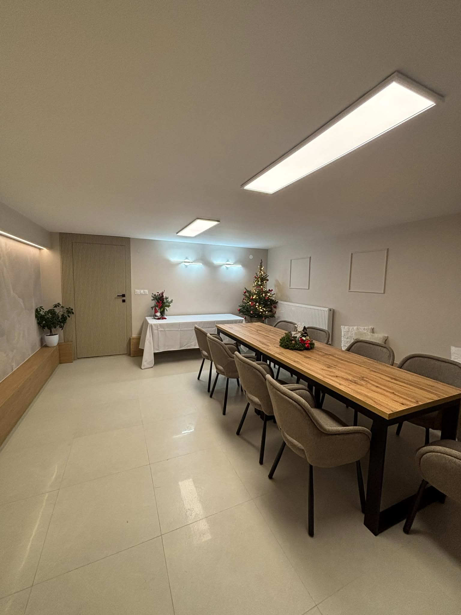 Jasne wnętrze z drewnianym stołem i krzesłami. Ściany w odcieniach beżu, podłoga wyłożona jasnymi płytkami. Dekoracje świąteczne dodają przytulnego charakteru.