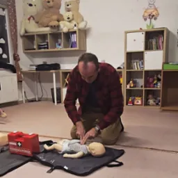 Instruktor demonstruje resuscytację krążeniowo-oddechową na manekinie niemowlęcia podczas kursu pierwszej pomocy. Czerwona apteczka obok. Dekoracje dziecięce w tle.