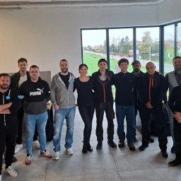 BHP CONCEPT JOANNA ANNA KOSKA - Grupa osób, prawdopodobnie uczestników szkolenia, pozuje do zdjęcia w jasnym pomieszczeniu z dużym oknem. Niektórzy mają na sobie odzież roboczą z logo firmy.