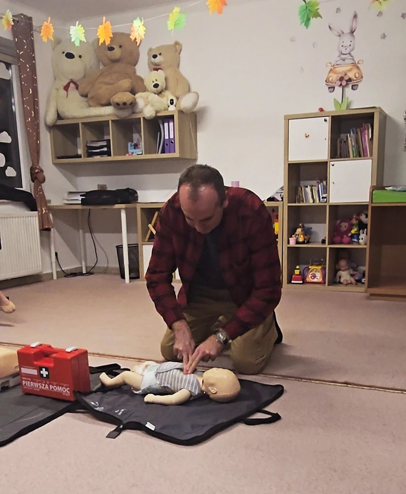 Instruktor demonstruje resuscytację krążeniowo-oddechową na manekinie niemowlęcia podczas kursu pierwszej pomocy. Czerwona apteczka obok. Dekoracje dziecięce w tle.