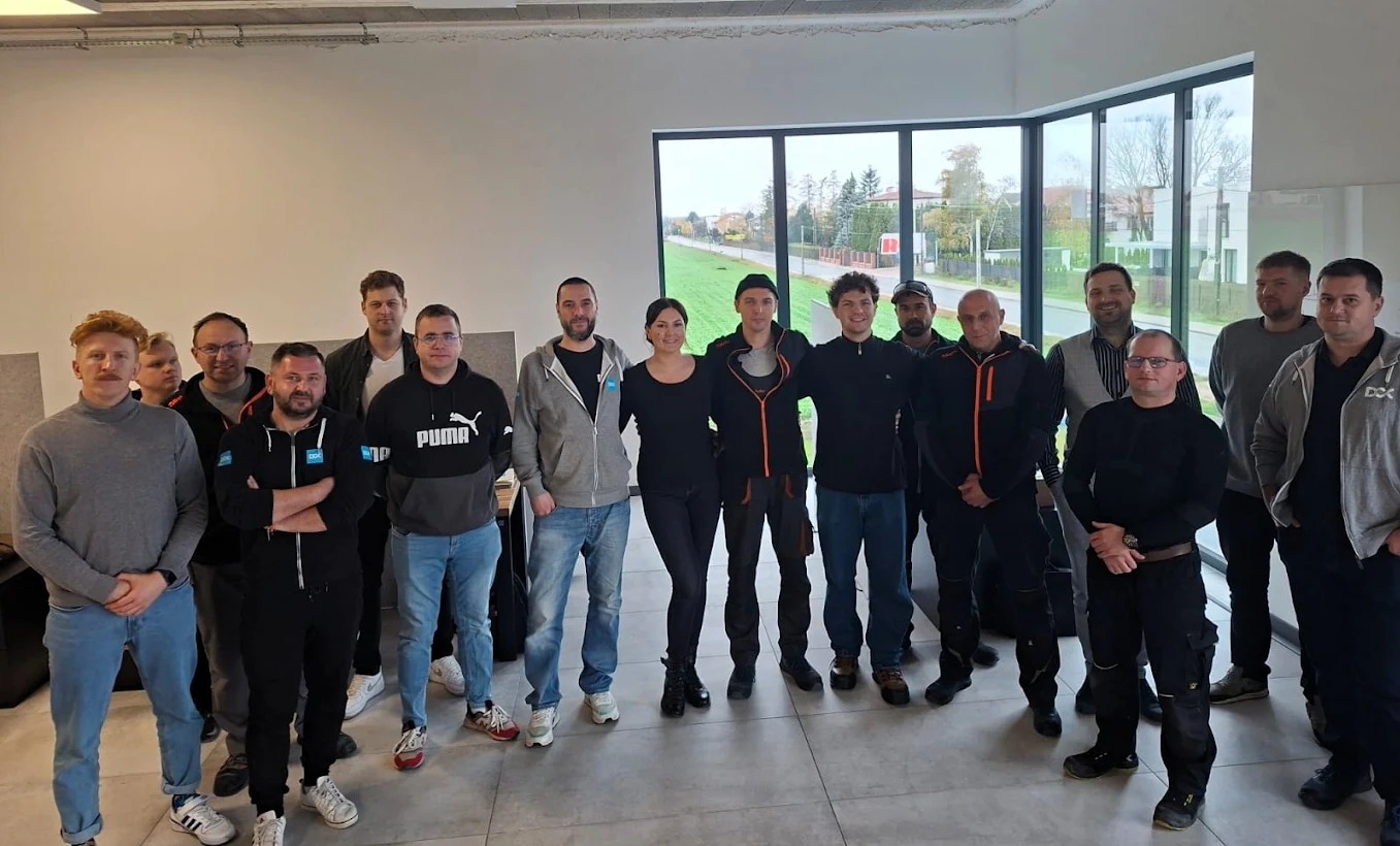 Grupa osób, prawdopodobnie uczestników szkolenia, pozuje do zdjęcia w jasnym pomieszczeniu z dużym oknem. Niektórzy mają na sobie odzież roboczą z logo firmy.