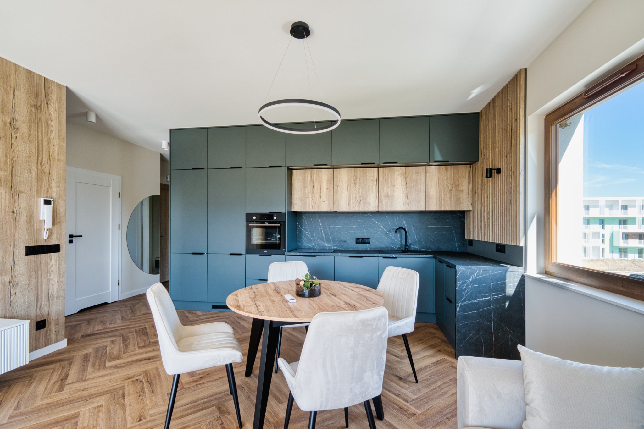 Nowoczesna kuchnia z okrągłym stołem i krzesłami. Zielone szafki, drewniane akcenty, parkiet w jodełkę. Minimalistyczna lampa wisząca. Jasne, przestronne wnętrze.