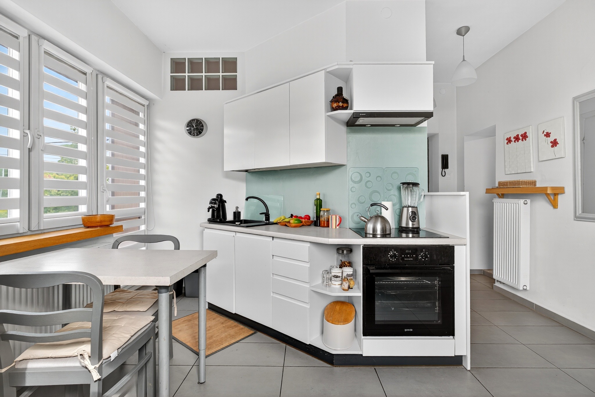 Jasna kuchnia w minimalistycznym stylu: białe meble, szare płytki, drewniane akcenty. Uwagę przykuwa okno z żaluzjami i dekoracje na ścianach.