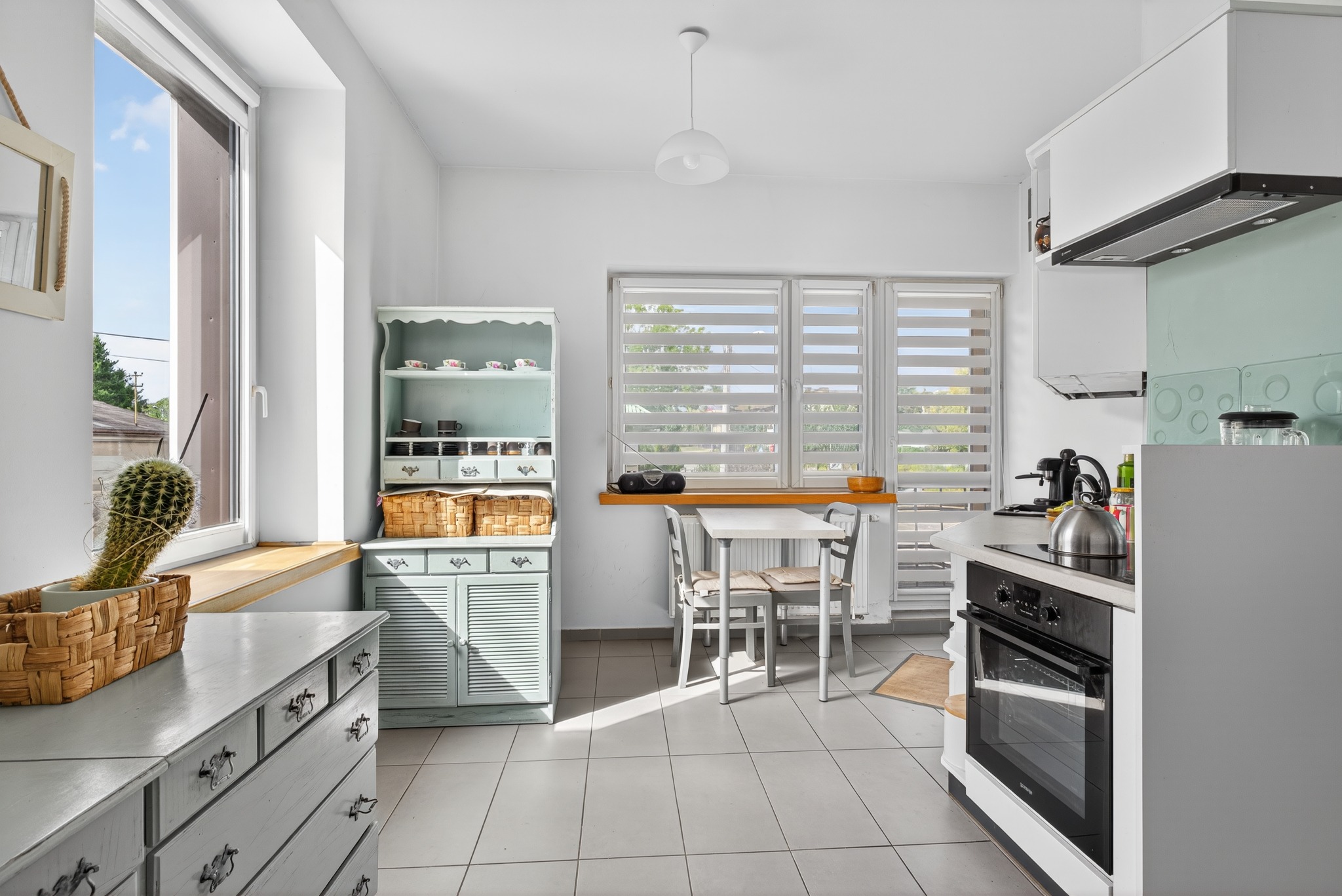 Jasna kuchnia w stylu vintage z błękitnymi meblami, białymi ścianami i kaktusem w oknie. Widok na stół z krzesłami i okno z żaluzjami.