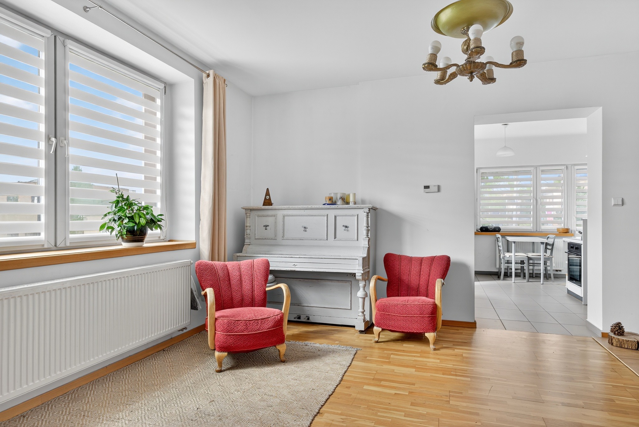 Jasne wnętrze z pianinem, dwoma czerwonymi fotelami i drewnianą podłogą. Widok na kuchnię przez otwarte drzwi. Roślina w oknie i ozdobny żyrandol.