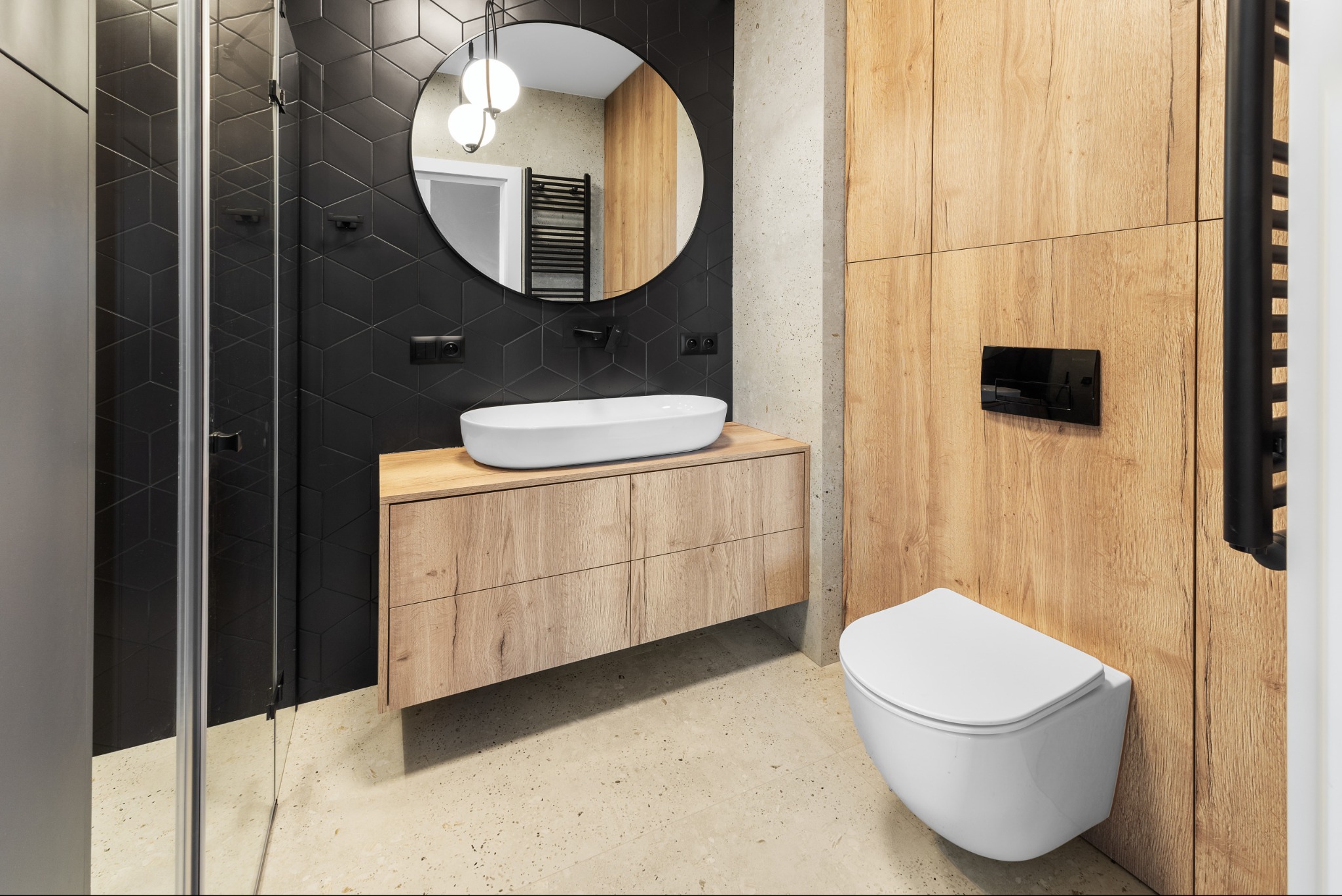Nowoczesna łazienka z czarnymi heksagonalnymi płytkami, drewnianą szafką pod umywalką i okrągłym lustrem z trzema kulami świetlnymi. Minimalistyczny design.