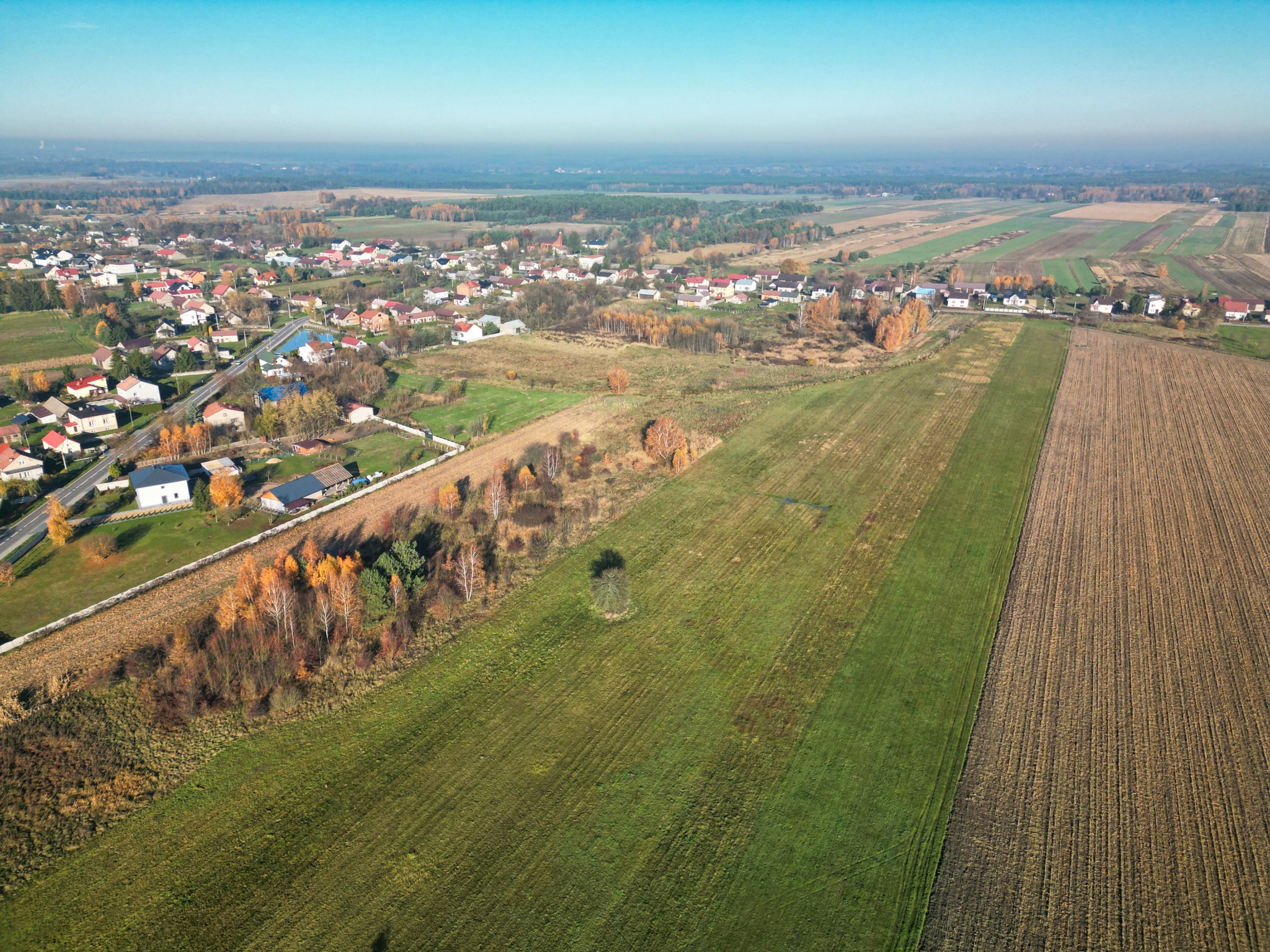 Panoramiczny widok z drona na wieś i pola uprawne w jesiennej scenerii. Widoczne domy z czerwonymi dachami, zielone i brązowe pola oraz drzewa w barwach jesieni.