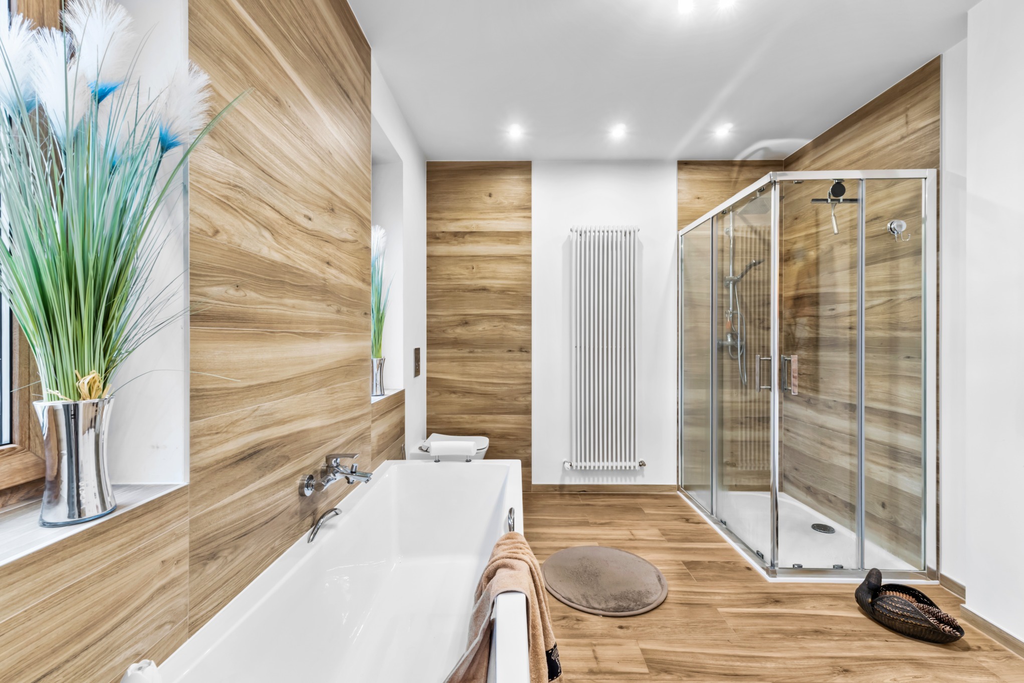 Nowoczesna łazienka z drewnianymi panelami, białą wanną i kabiną prysznicową. Dekoracyjny wazon z roślinami w oknie. Ciepłe oświetlenie podkreśla naturalny charakter wnętrza.