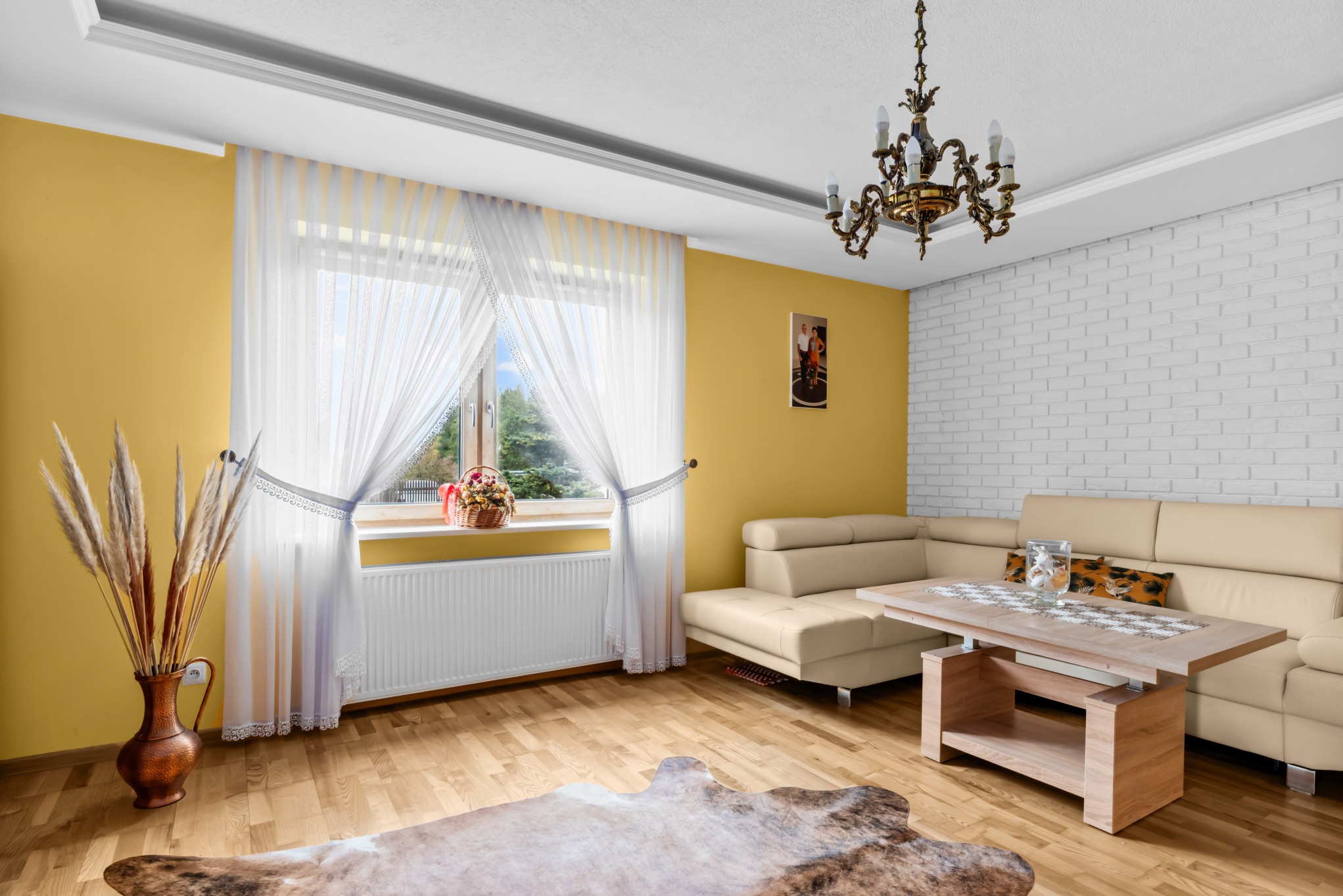 Jasny salon z żółtą ścianą, białym ceglanym fragmentem, beżową sofą i drewnianym stołem. W oknie widać firanki i kwiaty. Na podłodze skóra.
