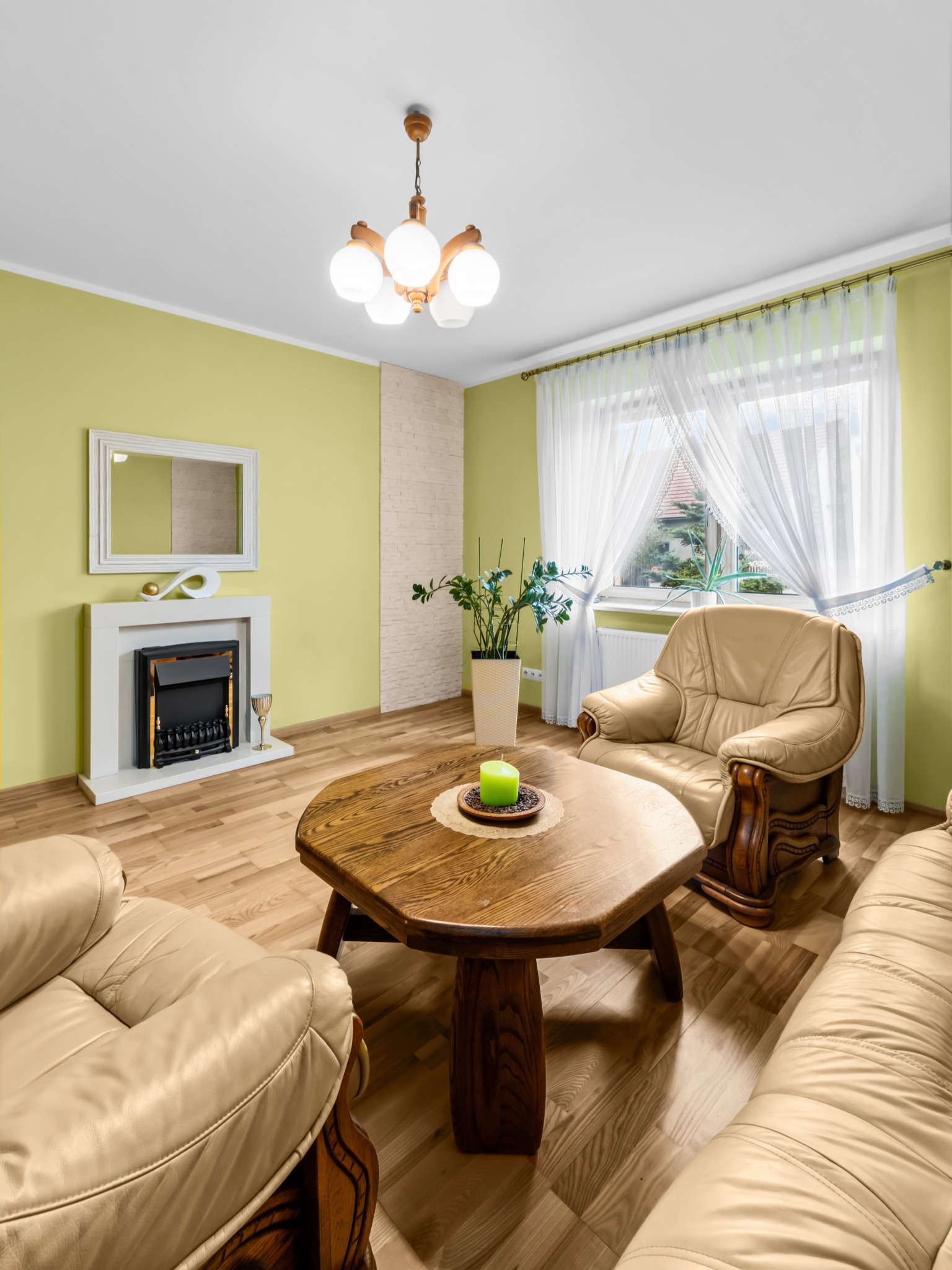 Jasne wnętrze salonu z beżowymi meblami skórzanymi, drewnianym stolikiem i kominkiem. Ściany w odcieniach zieleni, okno z białymi firankami. Widok na zewnątrz.