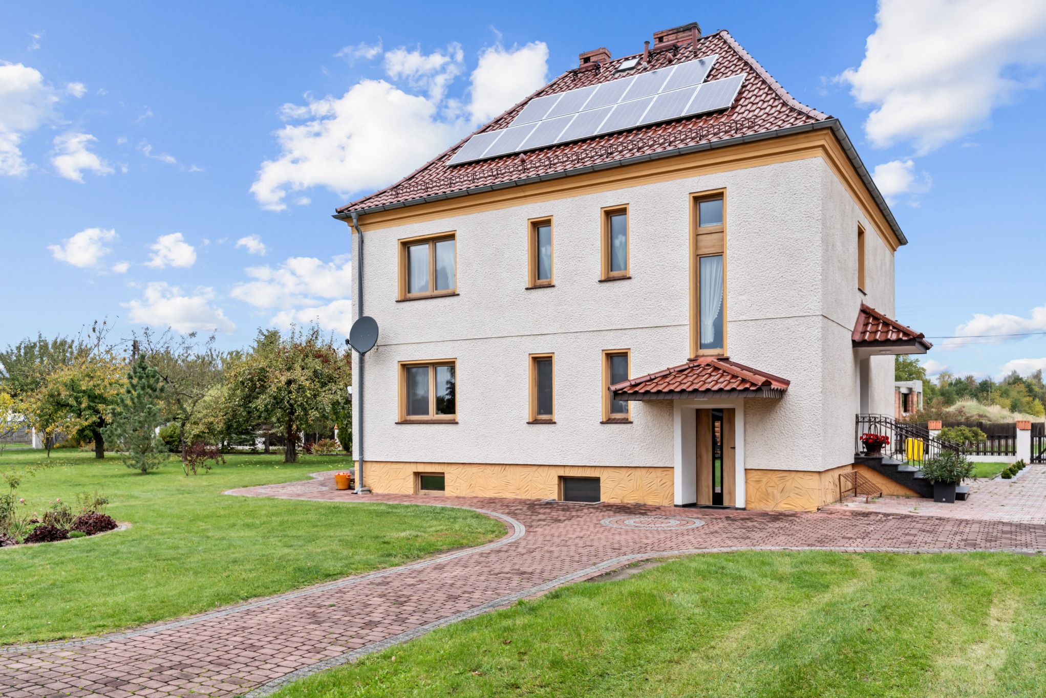 Dwupiętrowy dom z panelami słonecznymi na dachu, otoczony zielonym trawnikiem i ścieżką z czerwonej kostki. Widoczne okna z drewnianymi ramami i satelita na ścianie.