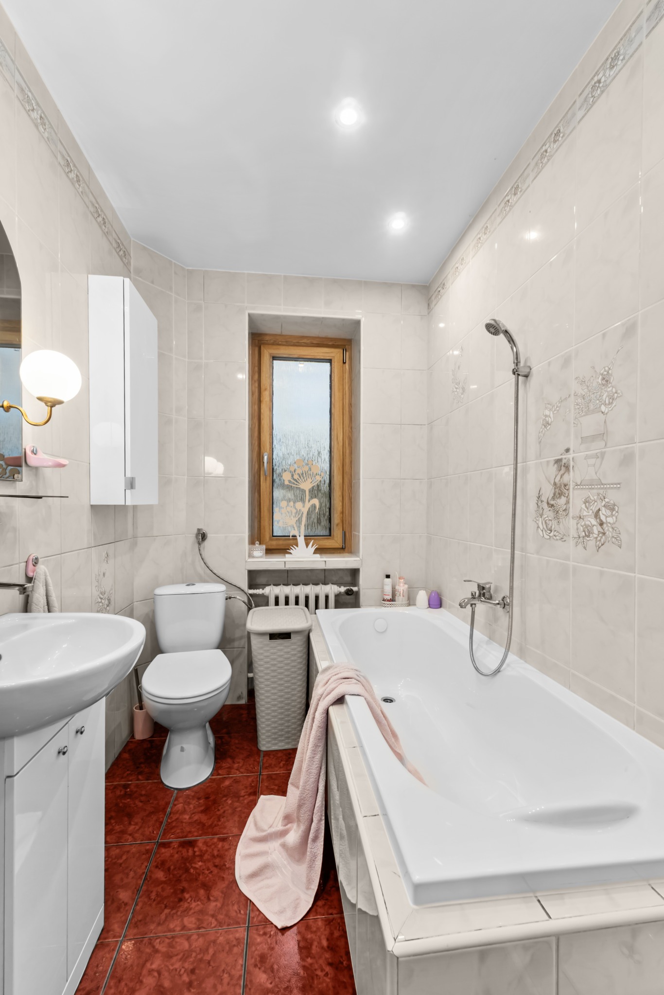 Jasna łazienka z wanną, umywalką i toaletą. Czerwona podłoga z płytek, beżowe ściany z dekoracyjnymi wzorami. Okno z drewnianą ramą. Ręcznik na wannie.