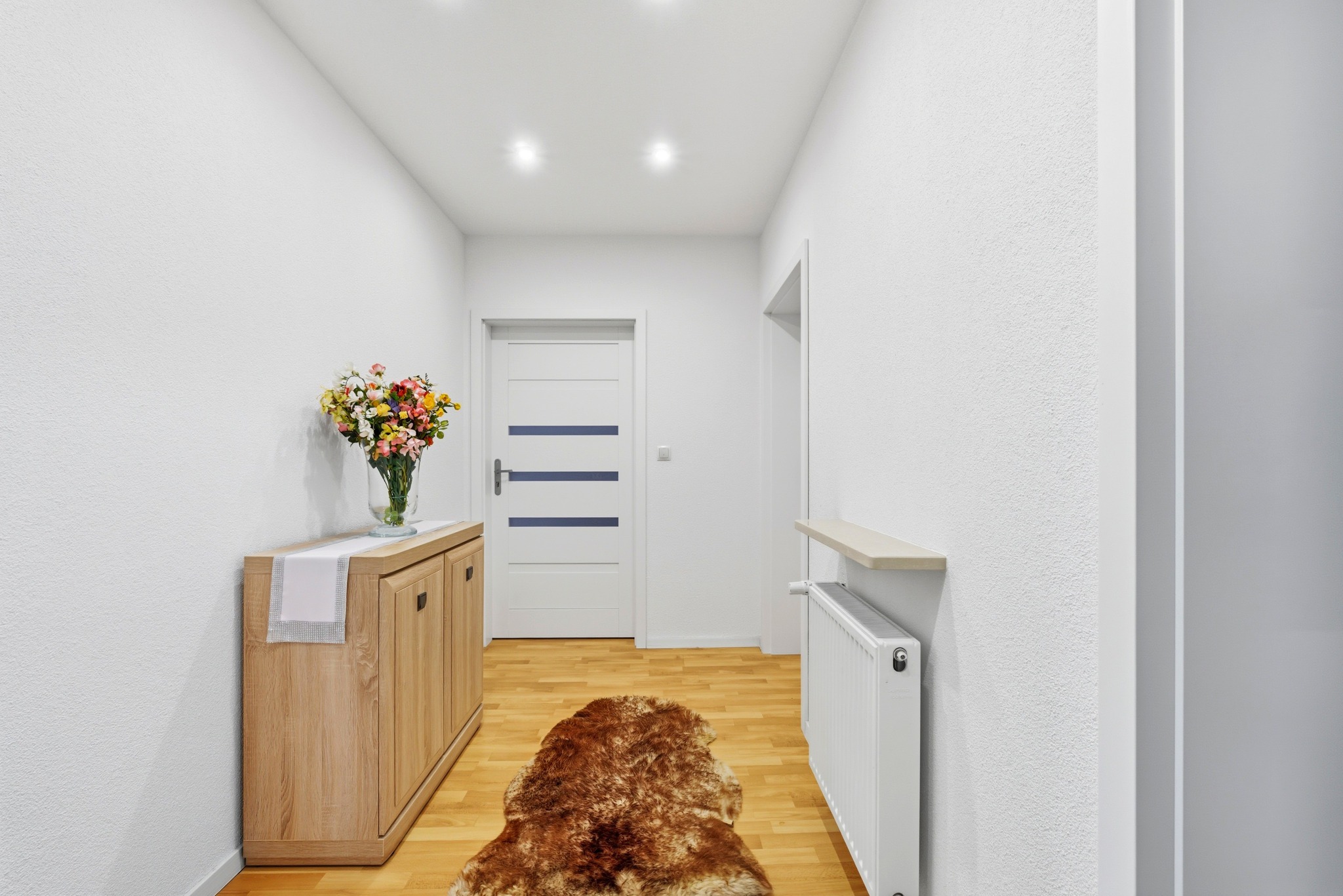 Jasny przedpokój z drewnianą komodą, bukietem kwiatów i futrzanym dywanem. Białe ściany, drzwi z poziomymi paskami i grzejnik. Minimalistyczny design.