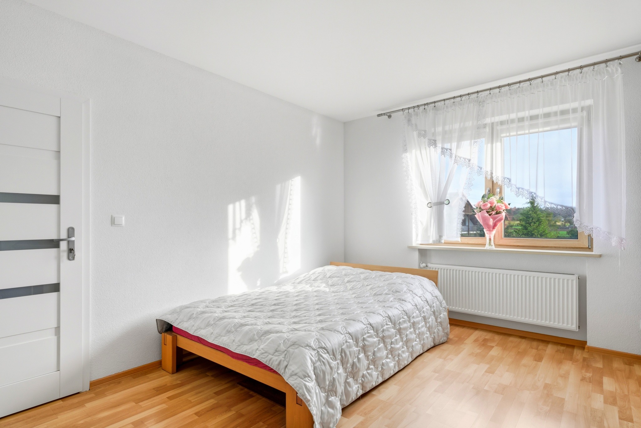 Jasna sypialnia w Częstochowie z drewnianą podłogą i łóżkiem. Białe ściany, okno z firankami i bukietem kwiatów. Minimalistyczny design.