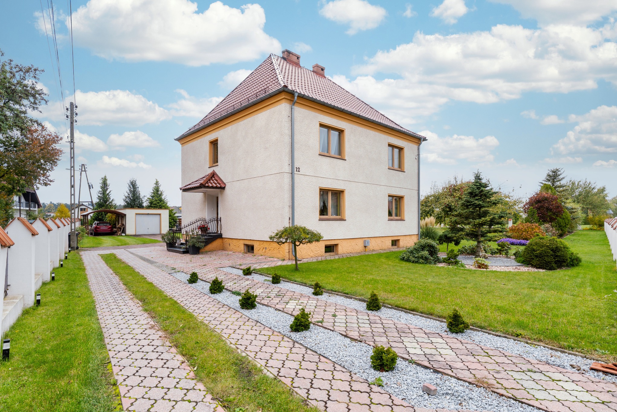Dwukondygnacyjny dom z beżową elewacją i czerwoną dachówką, otoczony zielonym ogrodem z brukowanym podjazdem i niskim żywopłotem. Słoneczny dzień, błękitne niebo z chmurami.