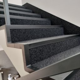 MONT - FLOOR Montaż PODŁÓG Marcin Rachel - Schody wyłożone ciemną wykładziną dywanową z szarymi listwami zabezpieczającymi krawędzie stopni. Widok z dołu, fragment balustrady w kadrze.
