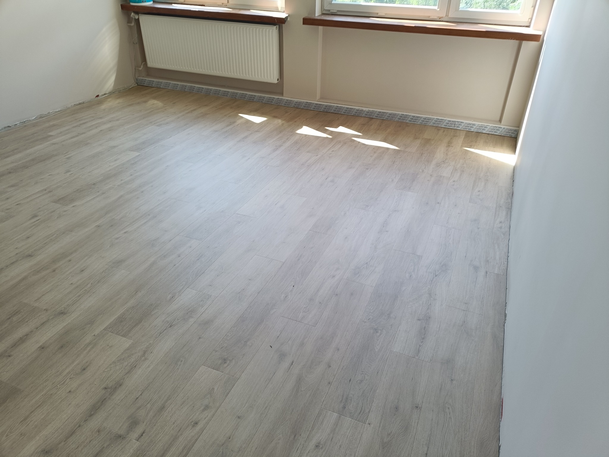 Jasne panele podłogowe ułożone w pokoju z białymi ścianami i oknami. Widoczne listwy przypodłogowe i grzejnik. Promienie słoneczne wpadają przez okno.