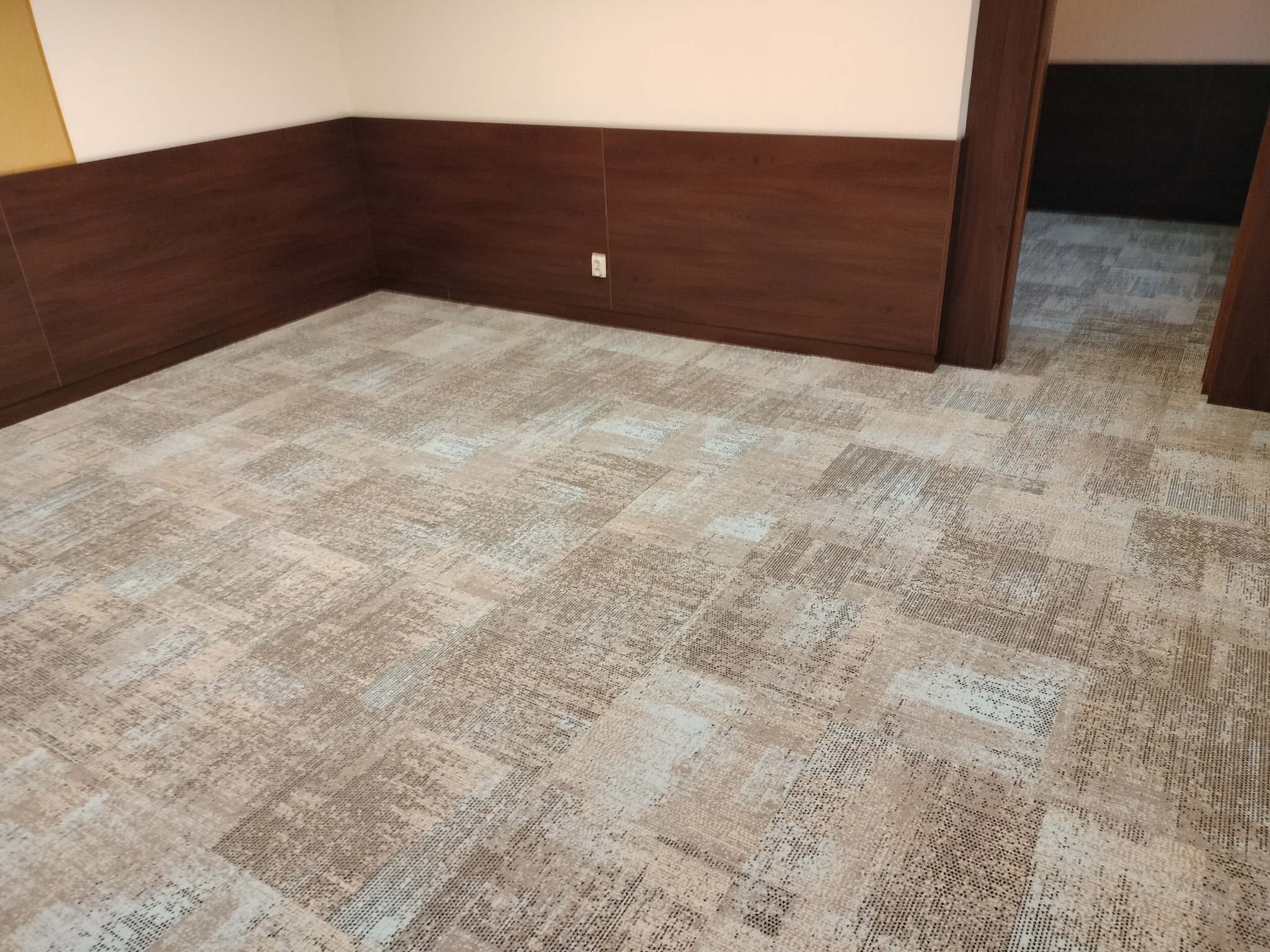 Wnętrze z nową, wzorzystą wykładziną podłogową w odcieniach beżu i brązu, dopasowaną do drewnianej boazerii. Widoczny fragment ściany i drzwi.