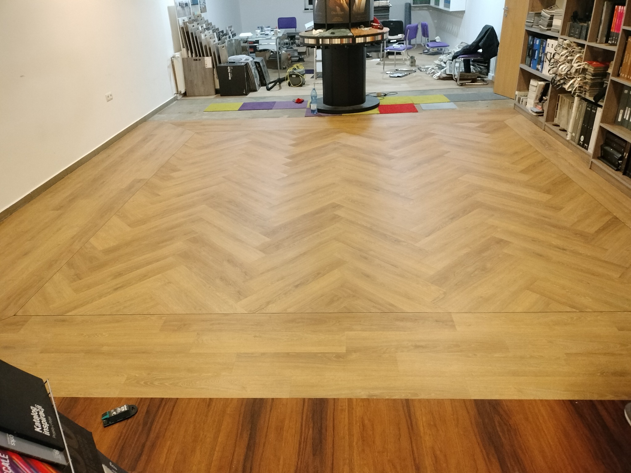 Jasne panele ułożone we wzór jodełki w przestronnym wnętrzu. Widoczne fragmenty ekspozycji paneli i regału z książkami. Kontrastowe obramowanie podłogi.