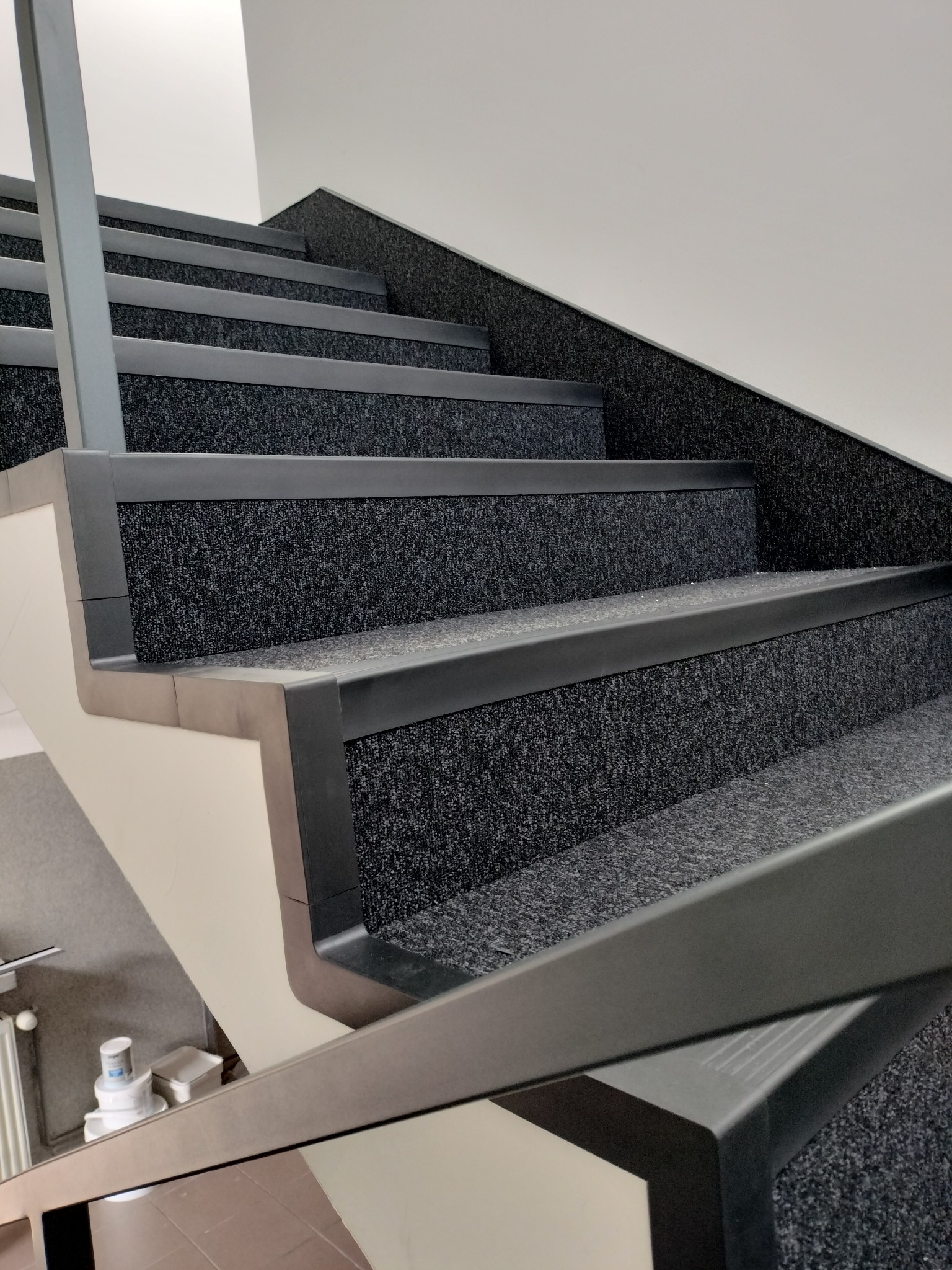 Schody wyłożone ciemną wykładziną dywanową z szarymi listwami zabezpieczającymi krawędzie stopni. Widok z dołu, fragment balustrady w kadrze.