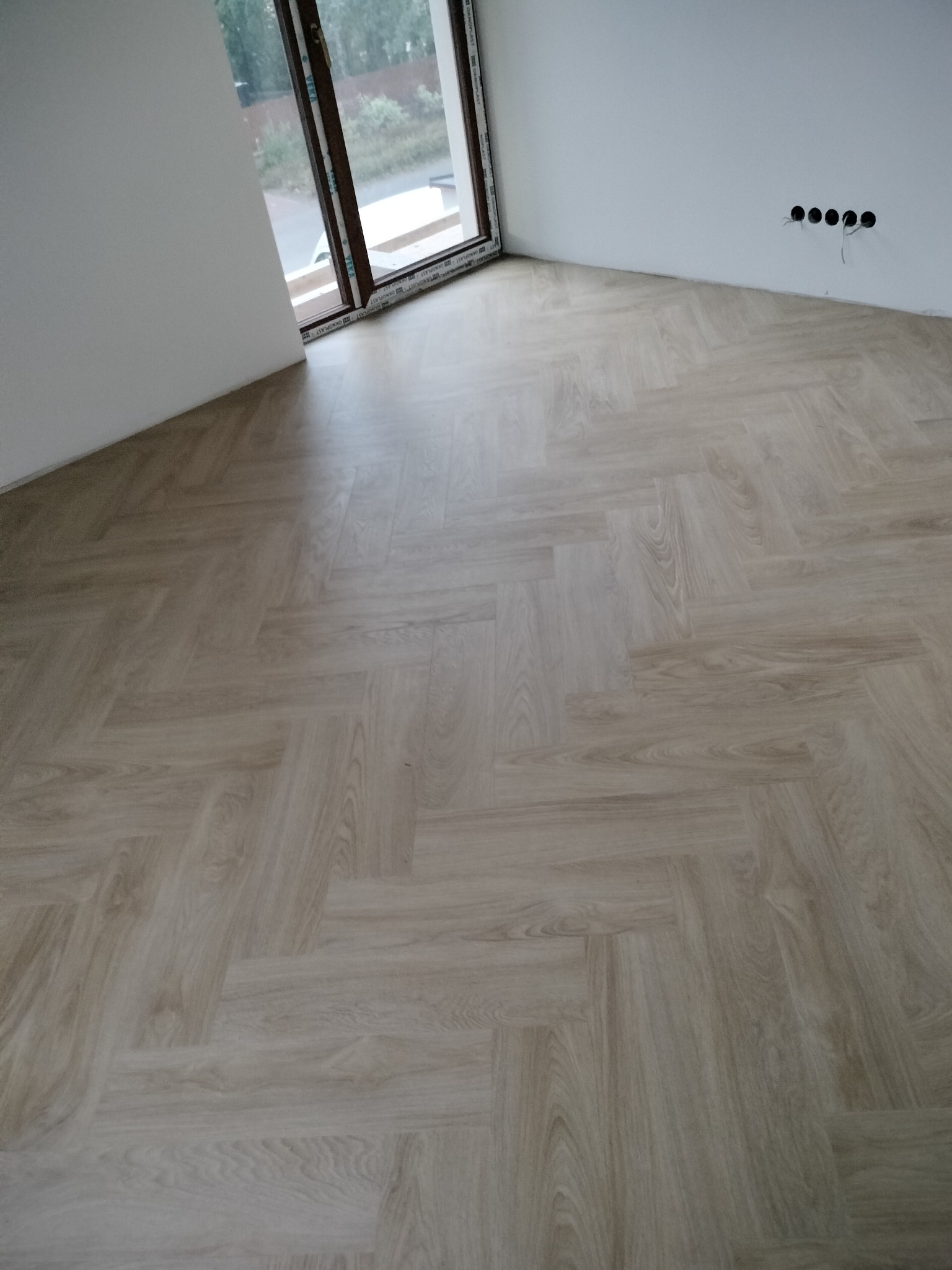 Jasne panele ułożone w jodełkę w nowym pokoju z oknem i gniazdkami elektrycznymi. Widok na podłogę po montażu, gotową do użytku.