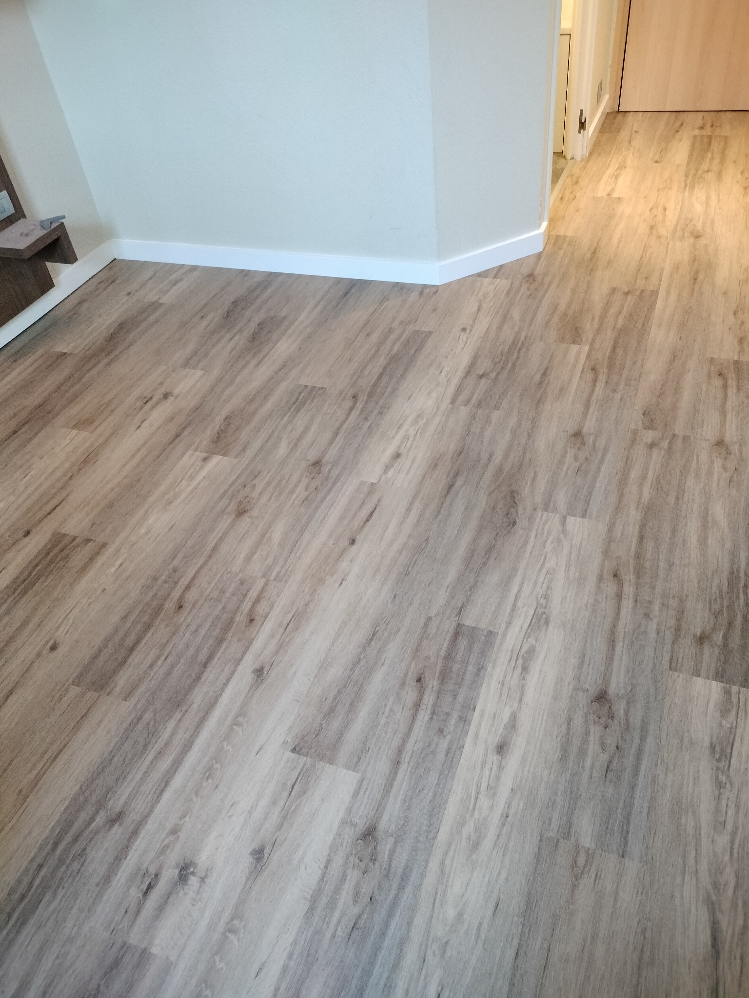 Jasne panele podłogowe z delikatnym wzorem drewna, ułożone wzdłuż pomieszczenia, z widocznymi białymi listwami przypodłogowymi i fragmentem drzwi w tle.