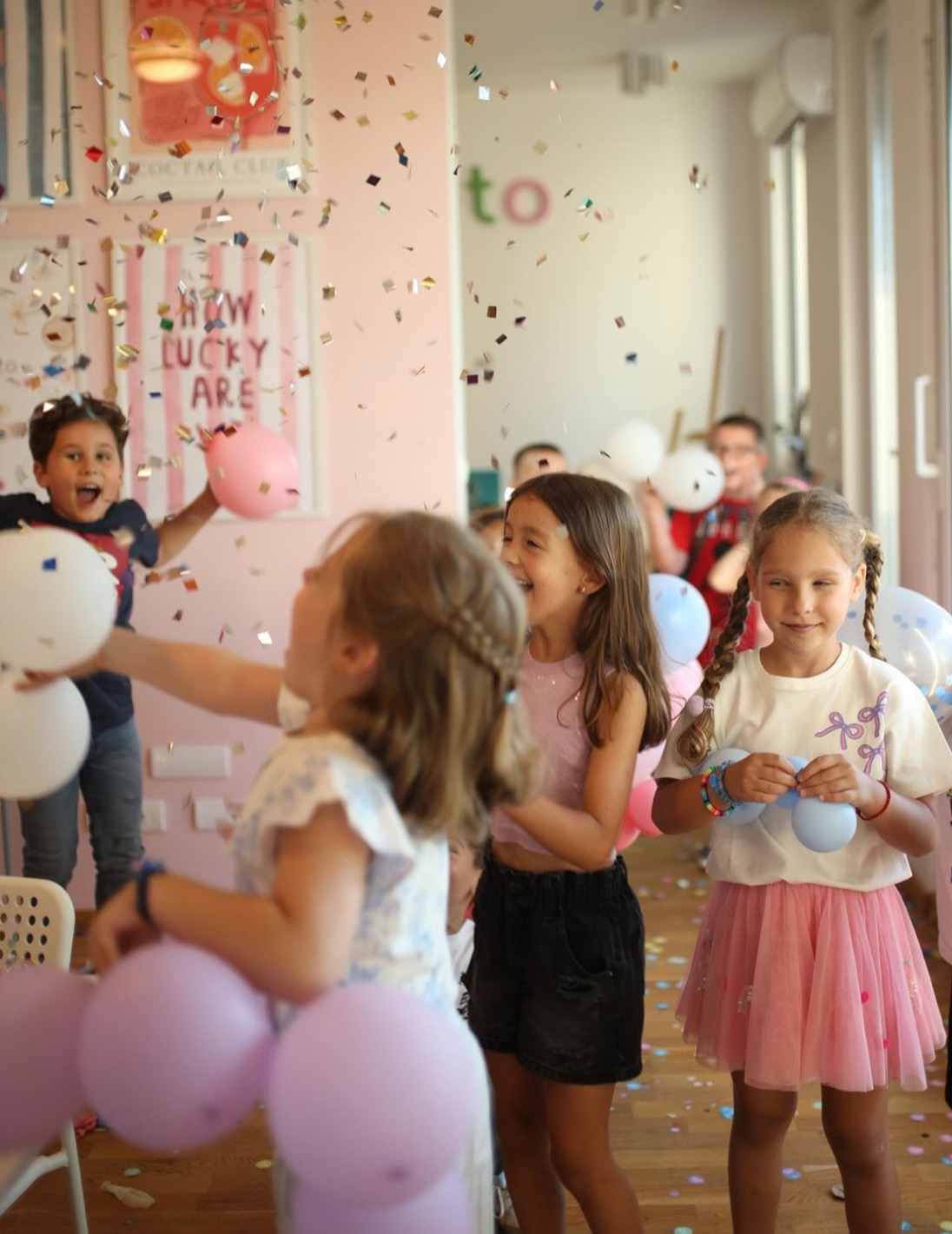 Uśmiechnięte dzieci w różowych strojach bawią się balonami podczas imprezy z konfetti w jasnym pomieszczeniu. Radosna atmosfera urodzinowa.