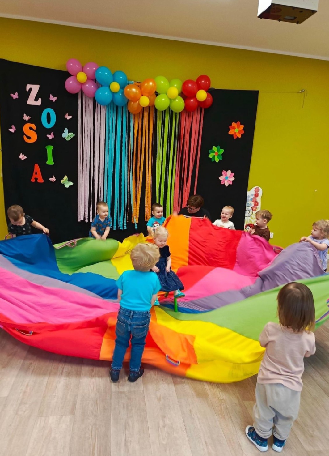 Animator prowadzi zabawy z kolorową chustą animacyjną dla grupy dzieci w sali z dekoracjami urodzinowymi. Widoczne balony i napis Zosia.