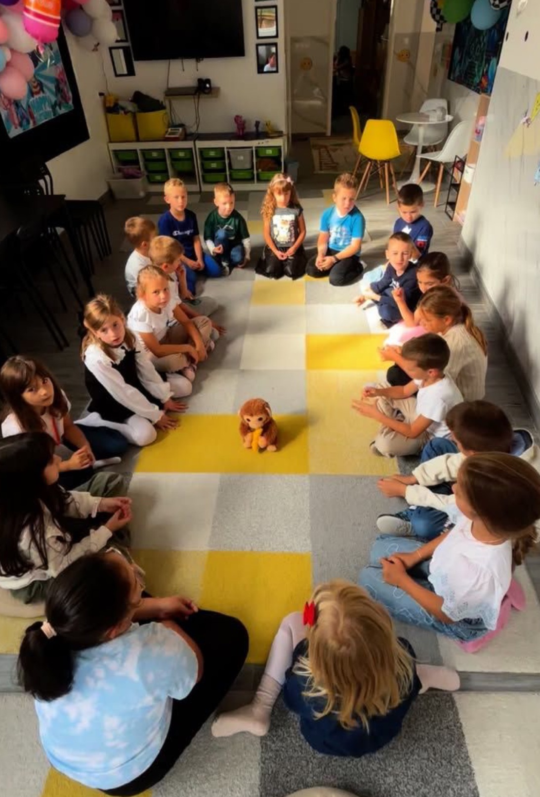 Dzieci siedzą w kręgu na podłodze w sali zabaw, skupione wokół pluszowej maskotki. Dekoracje balonowe i zabawki w tle sugerują imprezę urodzinową.