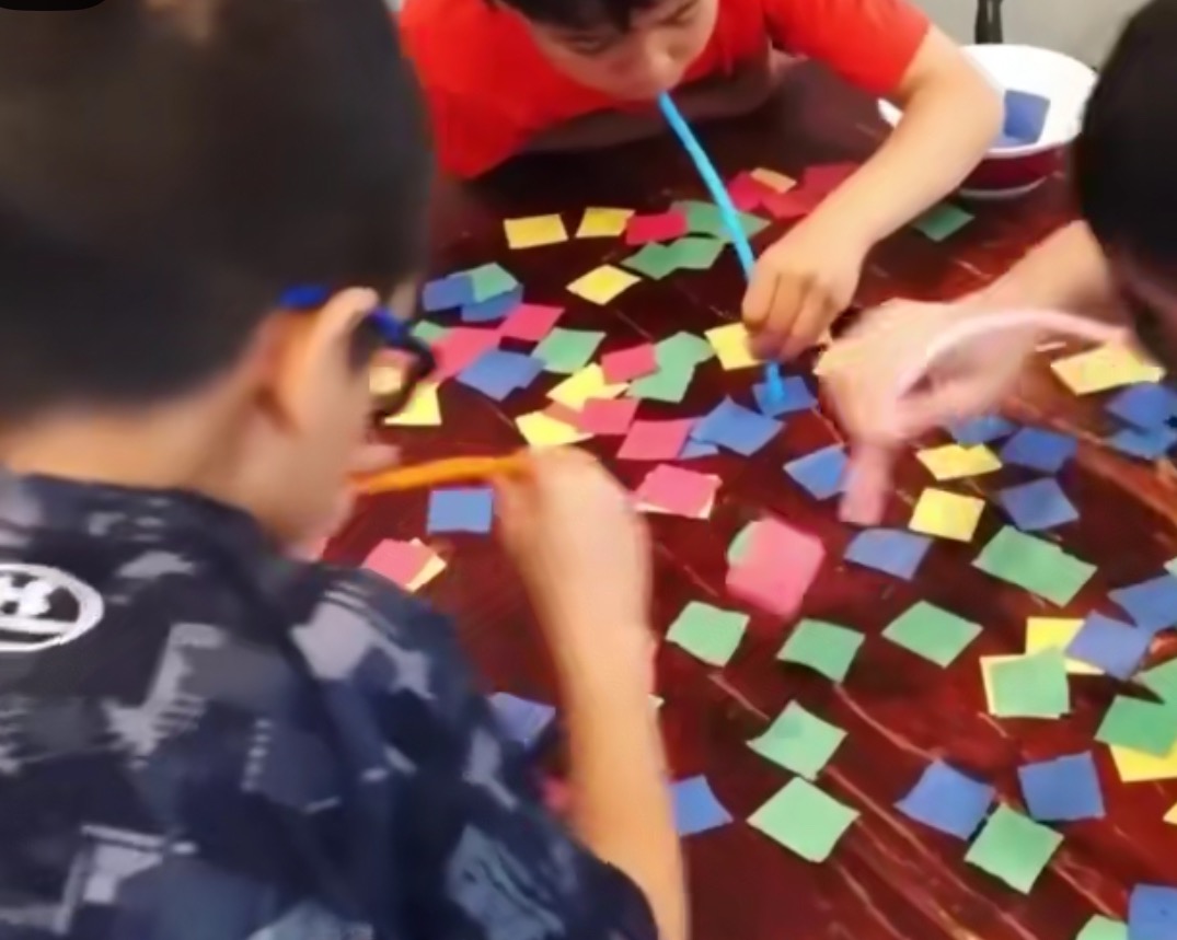 Dzieci rywalizują w grze z kolorowymi kwadratami i słomkami na stole. Zabawa edukacyjna, rozwijająca zręczność i koordynację wzrokowo-ruchową.