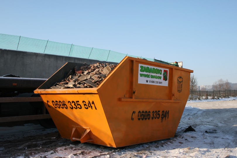 Pomarańczowy kontener wypełniony gruzem, z widocznym numerem telefonu i logo firmy Zarąbski Wywóz Odpadów, stoi na śniegu w słoneczny dzień.