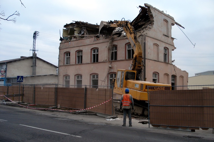 Żółta koparka CASE podczas rozbiórki trzypiętrowego budynku z cegły. Pracownik w kamizelce obserwuje proces. Częściowo zburzona konstrukcja, gruz.