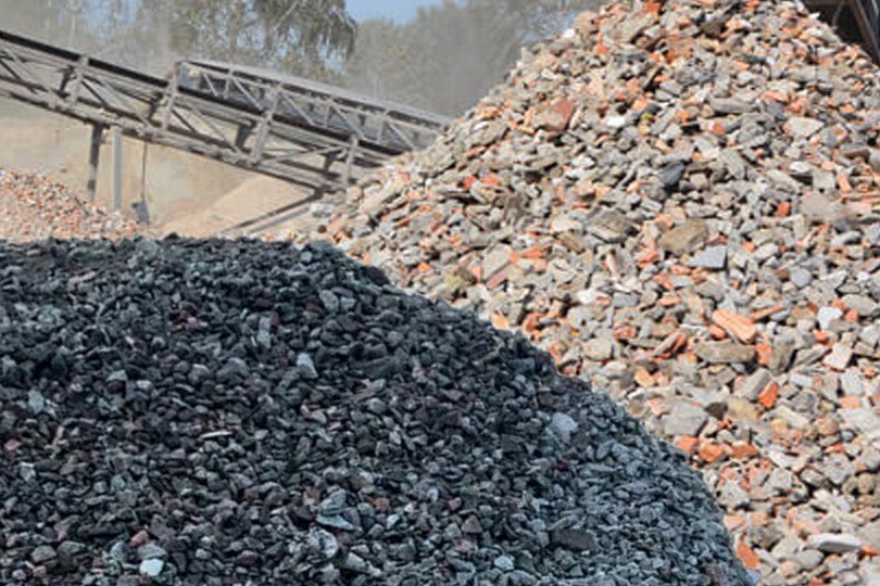 Dwa stosy gruzu: ciemny i jasny, z elementami cegieł, na tle konstrukcji taśmociągu. Widok z bliska, ukazujący strukturę materiałów.