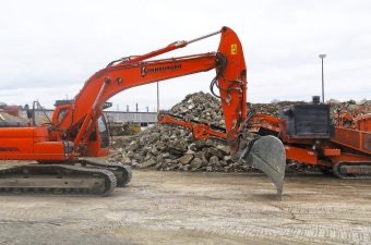 Pomarańczowa koparka z łyżką i kruszarka do gruzu na placu budowy, w tle sterta gruzu i pochmurne niebo. Widoczne detale maszyn i elementy konstrukcyjne.