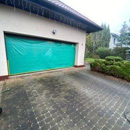 Przedsiębiorstwo Handlowo Usługowe Łukasz Muszyński - Przygotowanie do prac elewacyjnych: garaż zabezpieczony zieloną folią ochronną z żółtą taśmą, kostka brukowa przed budynkiem. Widoczne dekoracje świąteczne.