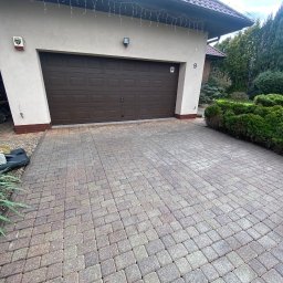 Przedsiębiorstwo Handlowo Usługowe Łukasz Muszyński - Dom z brązową bramą garażową i kostką brukową. Widoczny numer domu '9', alarm i dekoracyjne lampki. Otoczenie: krzewy i drzewa.