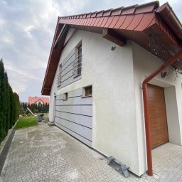 Przedsiębiorstwo Handlowo Usługowe Łukasz Muszyński - Nowoczesny dom jednorodzinny z elewacją w odcieniach bieli i szarości, z elementami drewnianymi i kostką brukową. Widok z perspektywy chodnika.