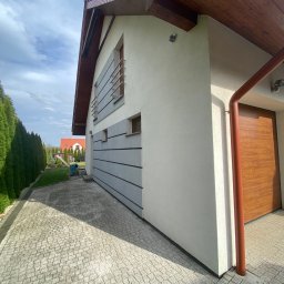 Przedsiębiorstwo Handlowo Usługowe Łukasz Muszyński - Nowoczesny dom z szarą elewacją i drewnianymi akcentami, widok z boku na kostkę brukową i zadbany ogród. Czyste linie i jasne kolory.