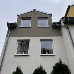 Przedsiębiorstwo Handlowo Usługowe Łukasz Muszyński - Dwukondygnacyjny budynek z szarą elewacją, widok z dołu. Widoczne okna z roślinami, rynny i trzy tuje przed budynkiem. Pochmurne niebo w tle.