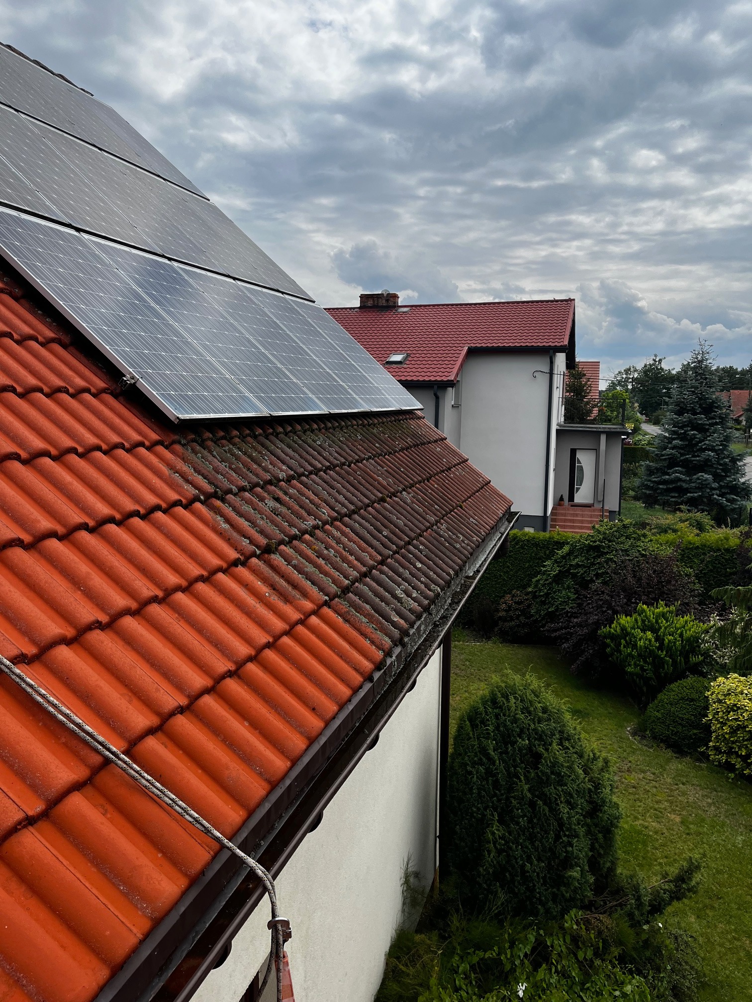 Częściowo umyty, czerwony dach z dachówki ceramicznej, z panelami słonecznymi w rogu. Widoczne zabrudzenia i mech na nieumytych fragmentach. W tle dom i zieleń.