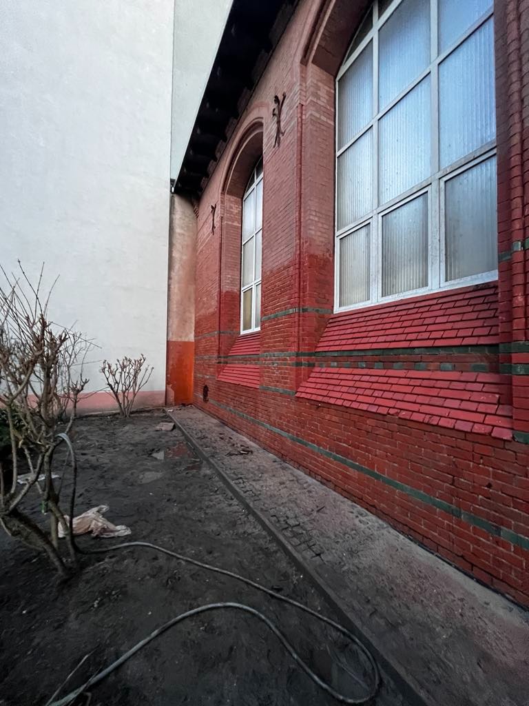 Ceglana elewacja budynku z dużymi oknami łukowymi, widoczna z perspektywy ziemi. Ślady po czyszczeniu/malowaniu, przewód na ziemi. Ujęcie z dołu.