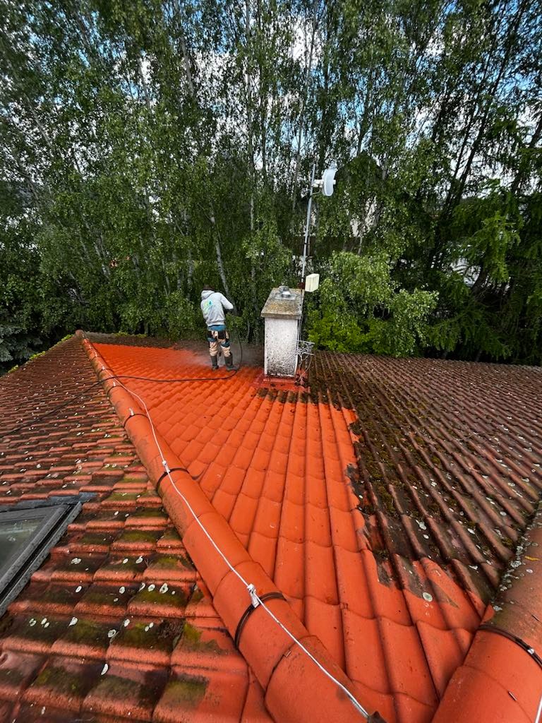 Częściowo umyty dach z czerwonej dachówki, widoczna różnica przed i po czyszczeniu. Pracownik w oddali, drzewa w tle. Antena na kominie.