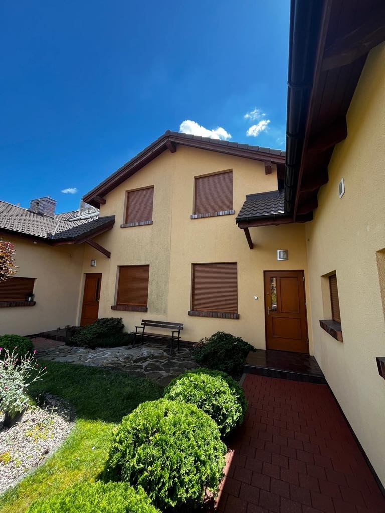 Jasna elewacja domu z brązowymi roletami i drzwiami, widok z poziomu ogrodu z zielonymi krzewami i czerwoną kostką brukową. Błękitne niebo z delikatnymi chmurami.