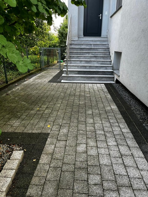 Częściowo wyczyszczona kostka brukowa prowadząca do schodów wejściowych budynku. Widoczne różnice w kolorze kostki przed i po czyszczeniu.