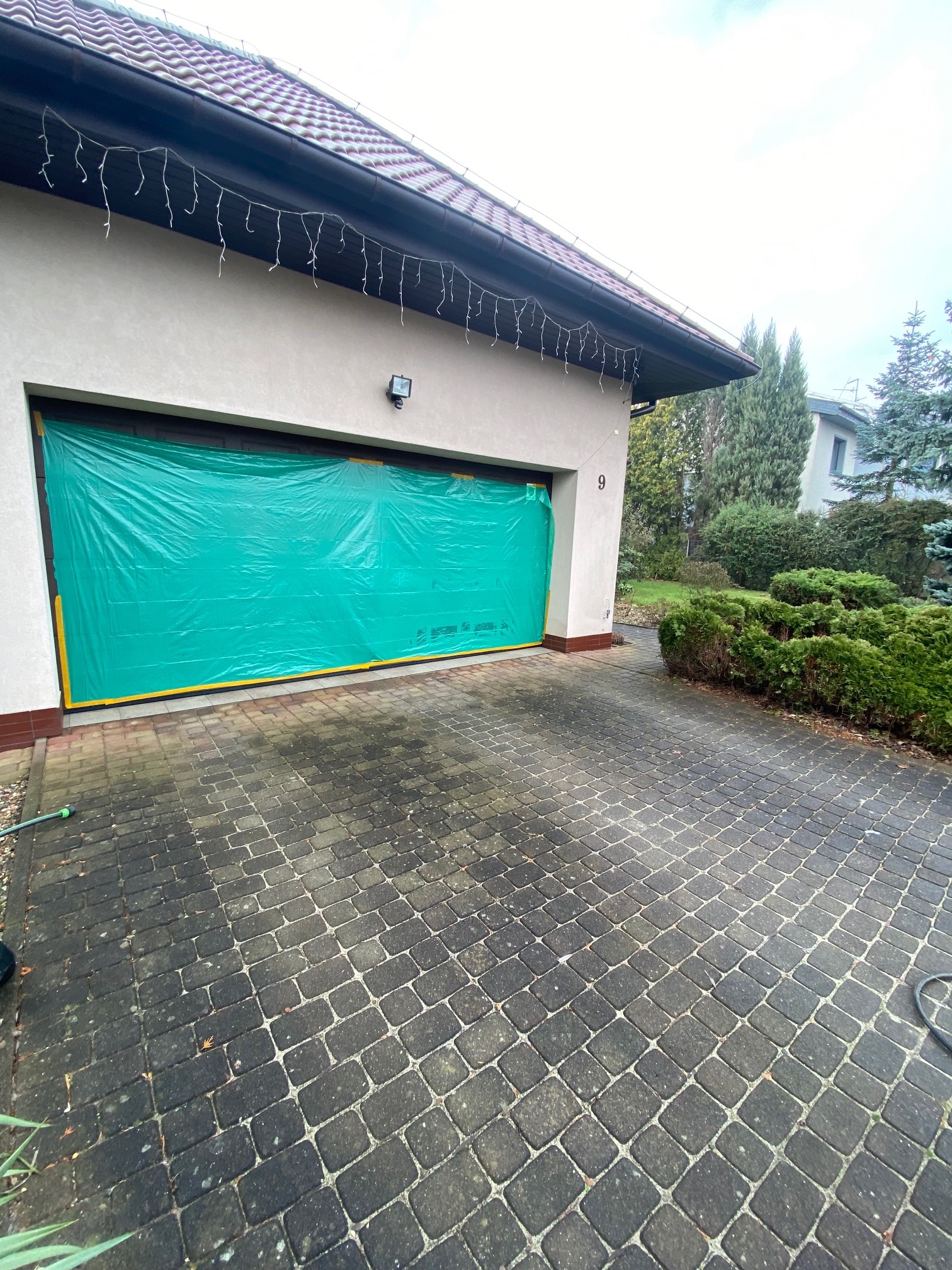 Przygotowanie do prac elewacyjnych: garaż zabezpieczony zieloną folią ochronną z żółtą taśmą, kostka brukowa przed budynkiem. Widoczne dekoracje świąteczne.