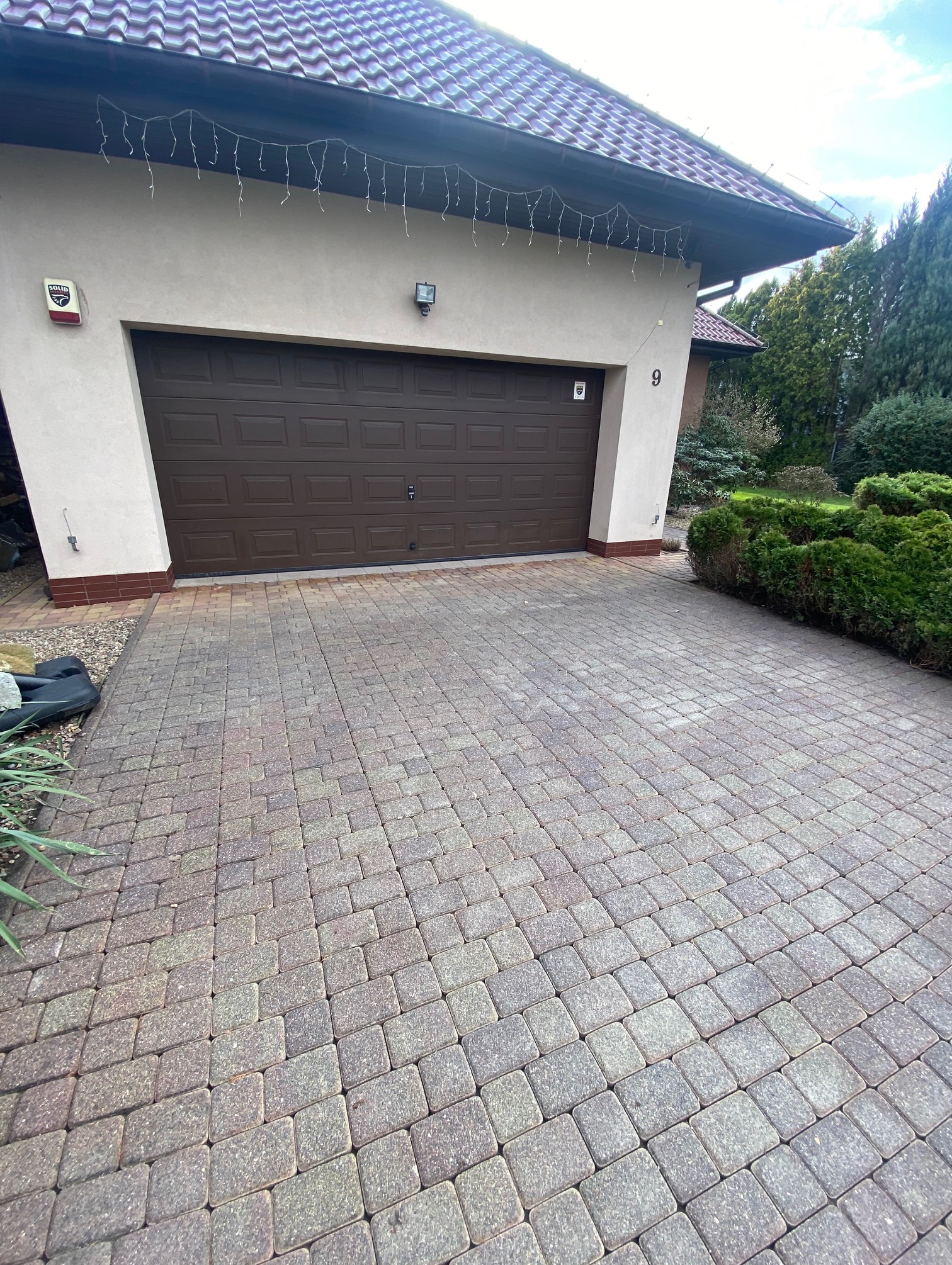 Dom z brązową bramą garażową i kostką brukową. Widoczny numer domu '9', alarm i dekoracyjne lampki. Otoczenie: krzewy i drzewa.