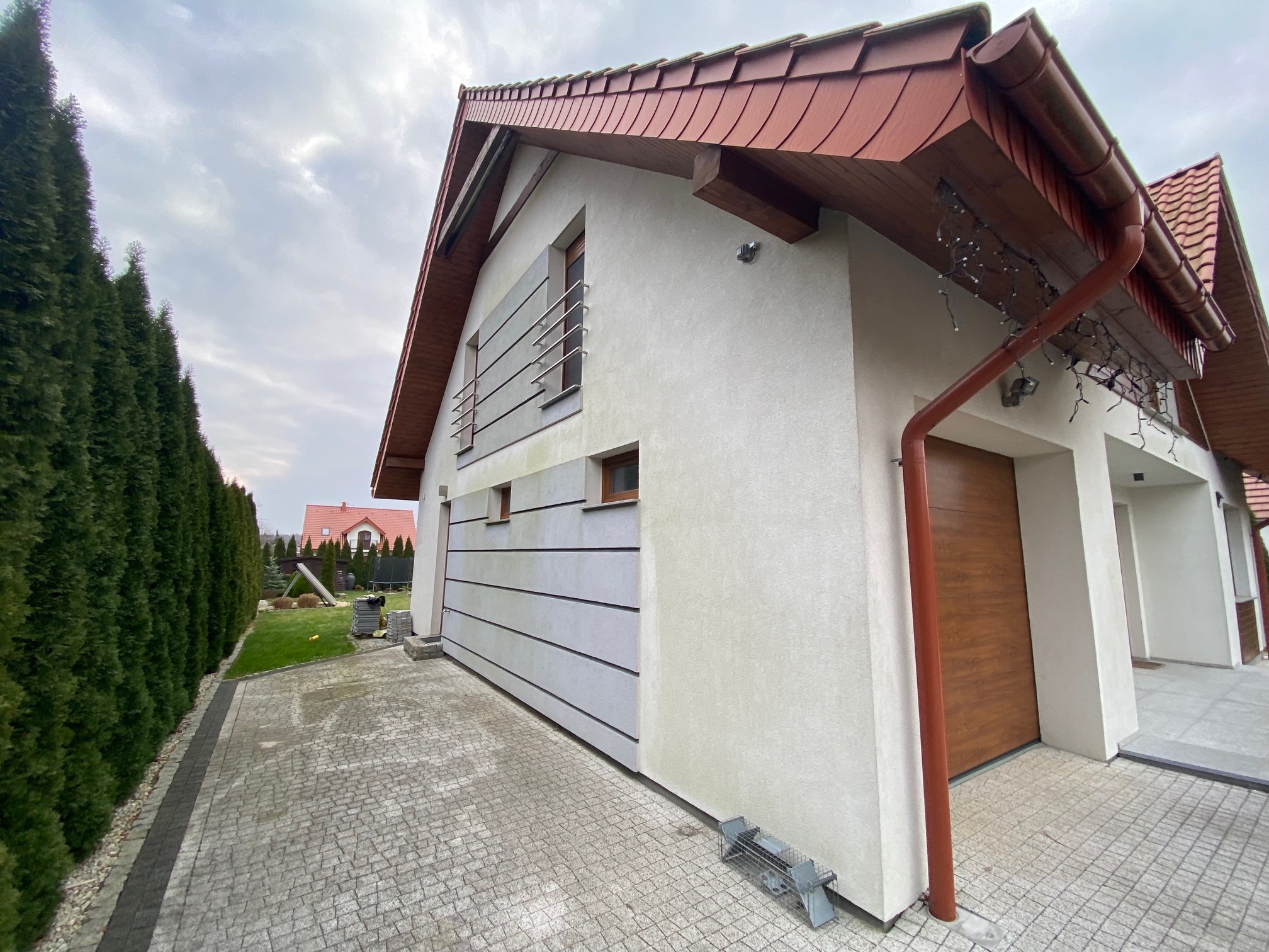 Nowoczesny dom jednorodzinny z elewacją w odcieniach bieli i szarości, z elementami drewnianymi i kostką brukową. Widok z perspektywy chodnika.