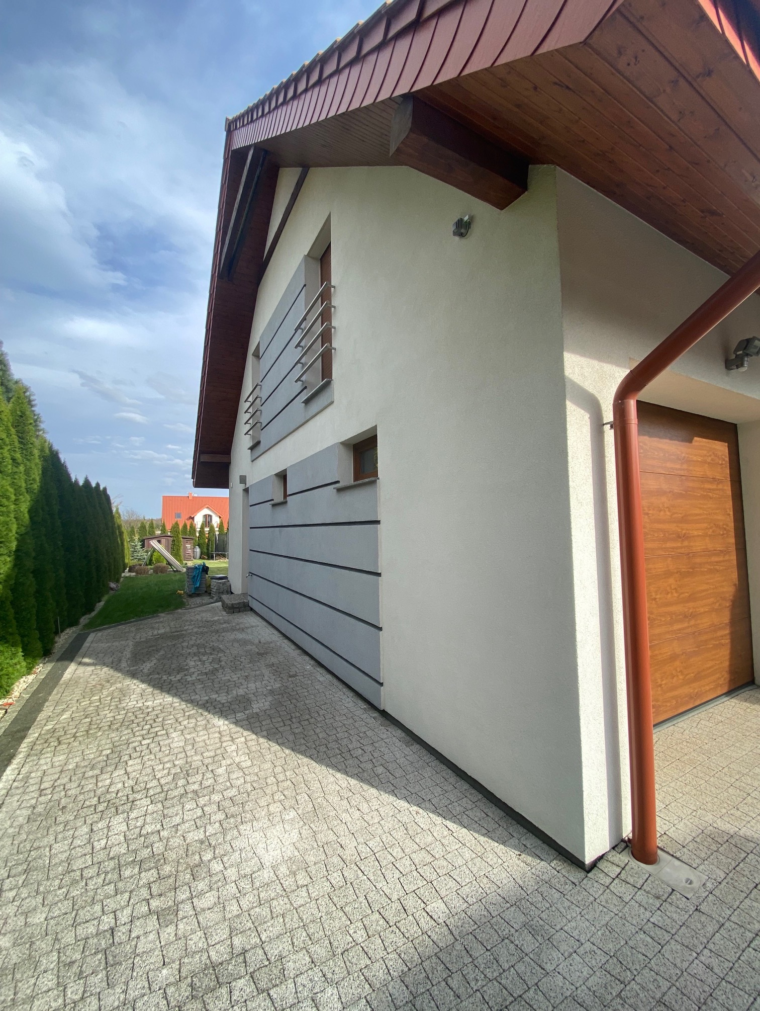 Nowoczesny dom z szarą elewacją i drewnianymi akcentami, widok z boku na kostkę brukową i zadbany ogród. Czyste linie i jasne kolory.